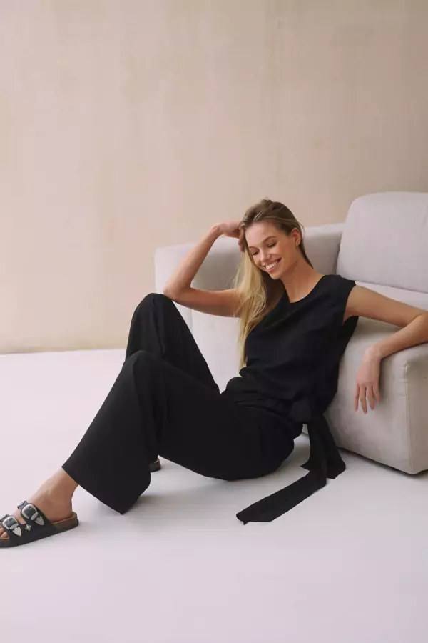 Czarna bluzka damska z krótkim rękawem, luźnym fasonem i wiązanym paskiem z boku, wykonana z wysokiej jakości materiału, na modelce stojącej oraz siedzącej na jasnym tle obok szarej kanapy.