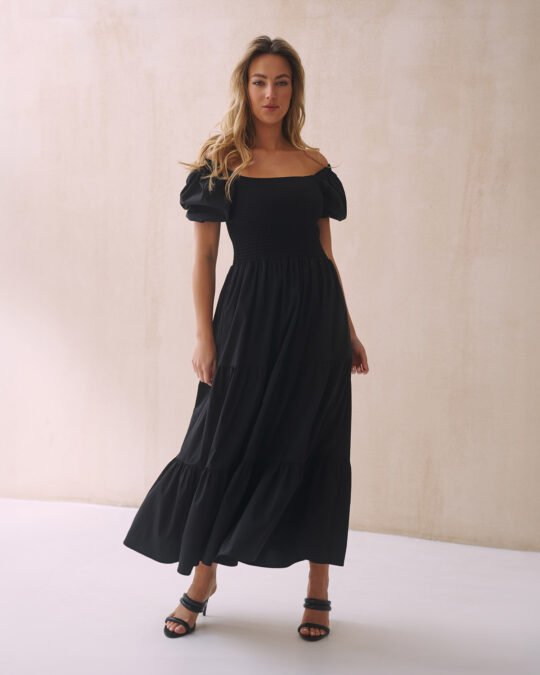Kobieta w czarnej sukience midi z bufiastymi rękawami i nicio-gumami na wysokości biustu, stojąca na jasnym tle. Sukienka idealnie dopasowuje się do sylwetki, podkreślając jej atuty, i jest wykonana z wysokiej jakości bawełny.