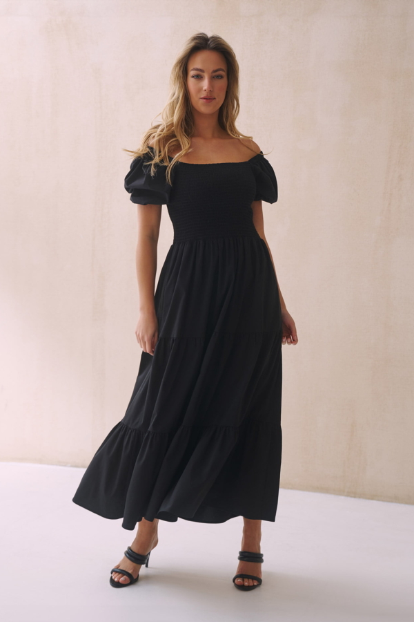 Kobieta w czarnej sukience midi z bufiastymi rękawami i nicio-gumami na wysokości biustu, stojąca na jasnym tle. Sukienka idealnie dopasowuje się do sylwetki, podkreślając jej atuty, i jest wykonana z wysokiej jakości bawełny.