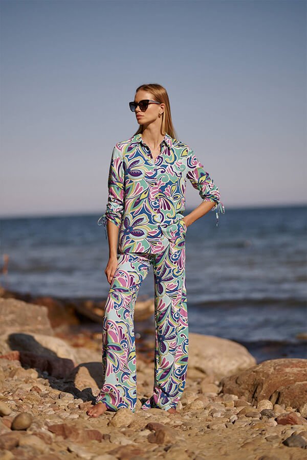 Kobieta w eleganckiej, kolorowej bluzce z kwiatowym wzorem, stojąca na tle morza i skalistej plaży.