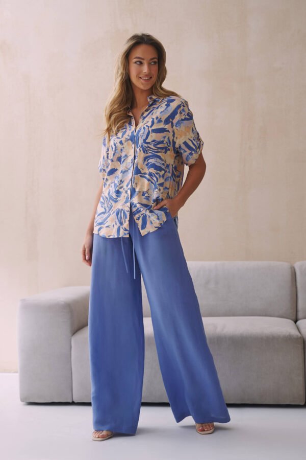 Kobieta w stylowej bluzce w odcieniach niebieskiego z abstrakcyjnym wzorem, zestawiona z szerokimi niebieskimi spodniami, pozująca w jasnym pomieszczeniu.