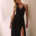 Kobieta w eleganckiej czarnej sukience wieczorowej z delikatną koronką na dekolcie i odkrytymi plecami, prezentująca rozcięcie z przodu eksponujące długie nogi.