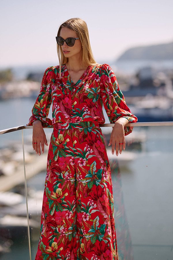 Kobieta ubrana w kolorową, tropikalną bluzkę z długimi rękawami, stojąca na tarasie z widokiem na port. Bluzka ma żywy, kwiatowy wzór w czerwonych, różowych i zielonych odcieniach. Kobieta nosi okulary przeciwsłoneczne i wygląda stylowo oraz elegancko w letnim otoczeniu.