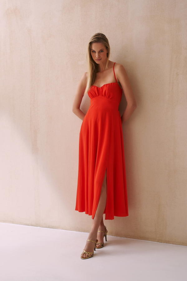 Kobieta ubrana w czerwoną sukienkę z rozcięciem z przodu, stojąca przy ścianie.