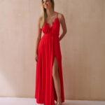 Kobieta w eleganckiej czerwonej sukience wieczorowej z delikatną koronką na dekolcie i odkrytymi plecami, prezentująca rozcięcie z przodu eksponujące długie nogi.