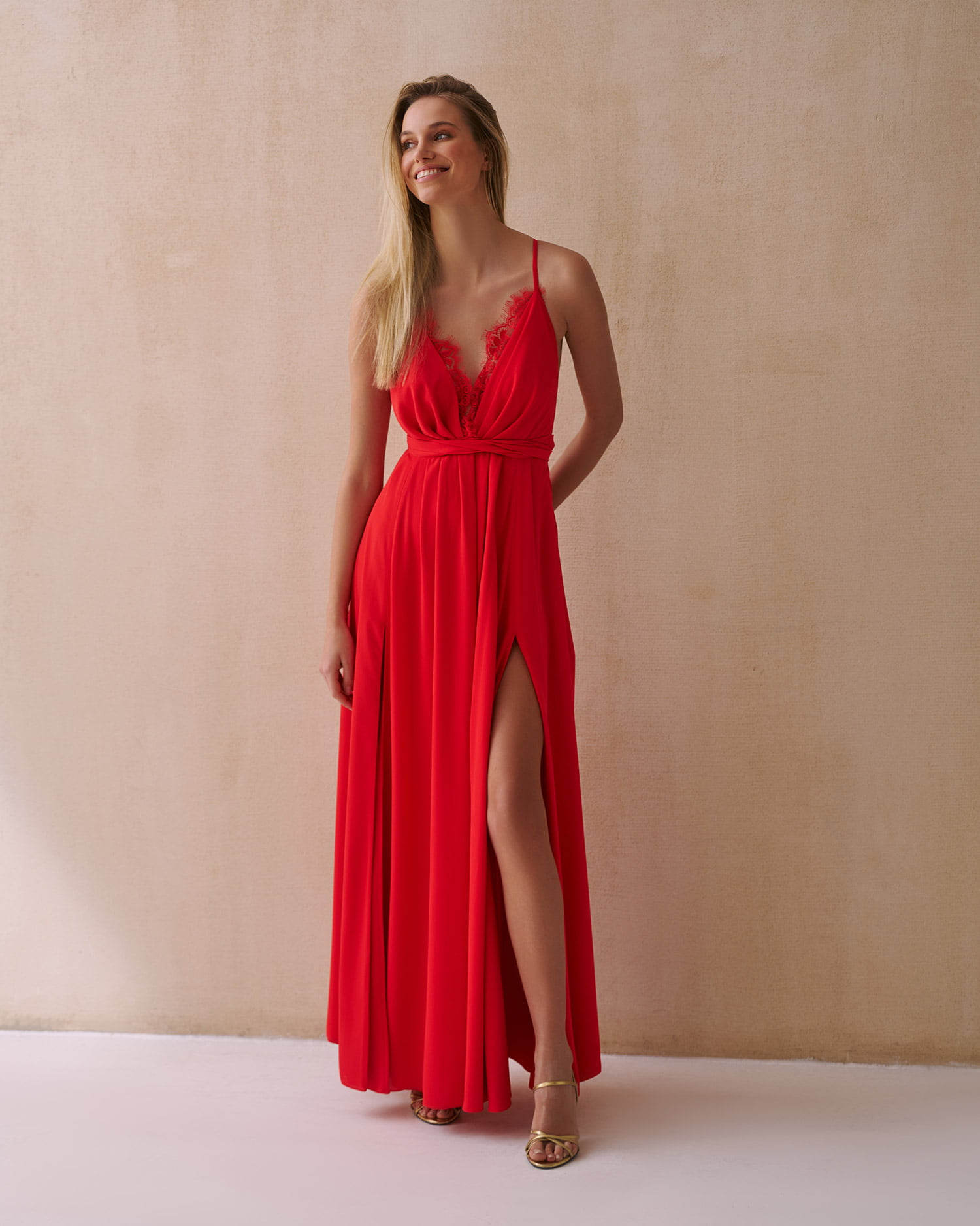 Kobieta w eleganckiej czerwonej sukience wieczorowej z delikatną koronką na dekolcie i odkrytymi plecami, prezentująca rozcięcie z przodu eksponujące długie nogi.