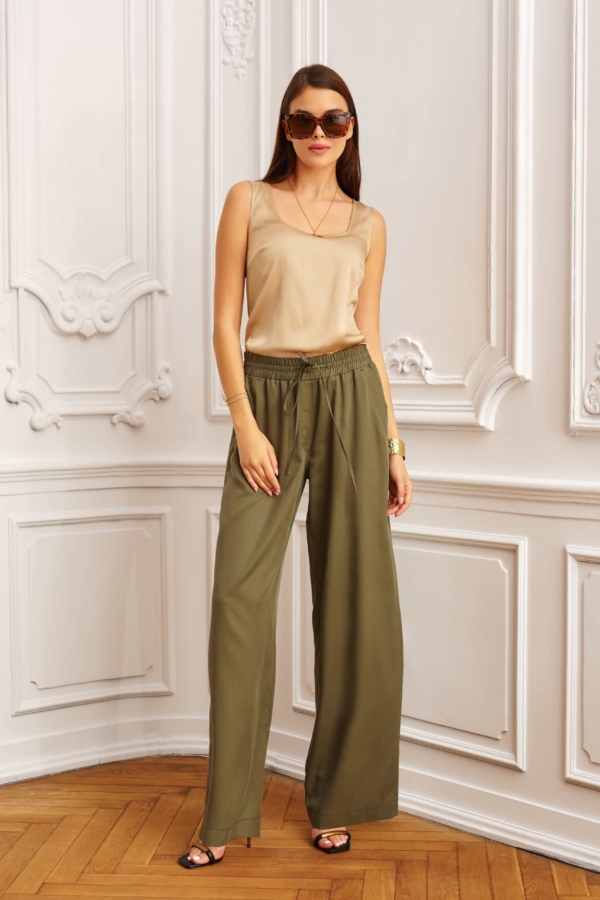 Modelka w khaki spodniach z szerokimi nogawkami i beżowym topie, pozująca w eleganckim wnętrzu