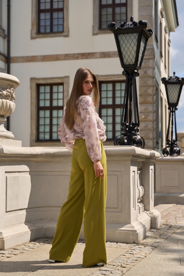 Modelka w eleganckich szerokich spodniach z wysokim stanem Milano w odcieniu zgaszonej limonki, z lekko rozszerzaną nogawką i gumką z tyłu pasa.