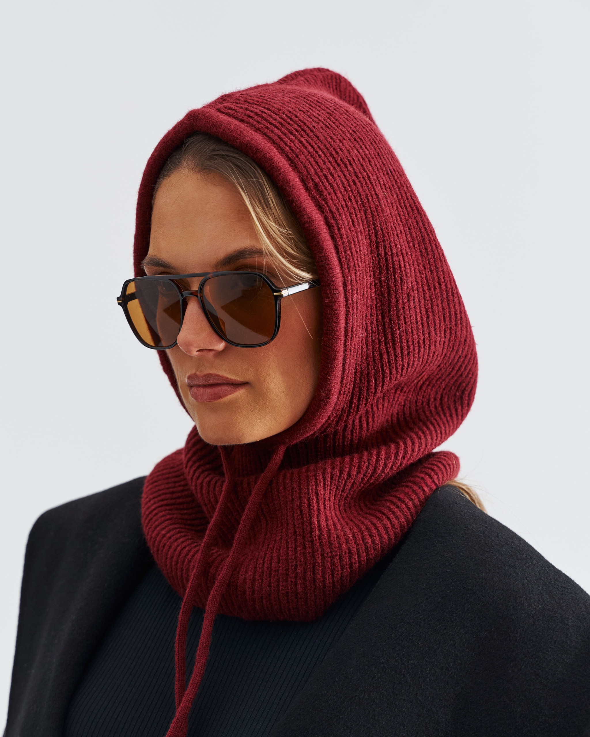 Modelka w bordowej dzianinowej balaclava, w okularach przeciwsłonecznych, w czarnym płaszczu – modna zimowa stylizacja.