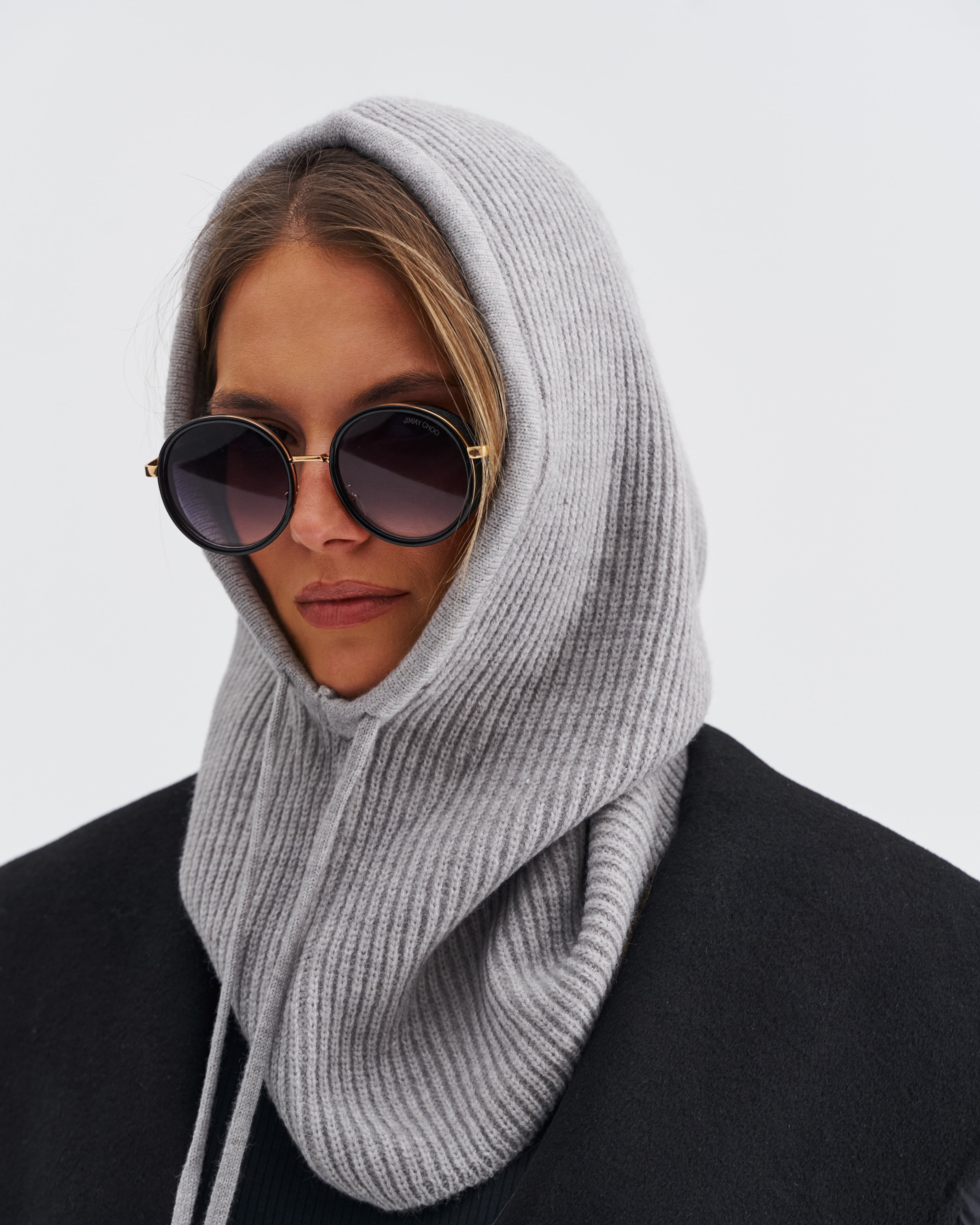 Modelka w szarej dzianinowej balaclava, w okularach przeciwsłonecznych, w połączeniu z czarnym płaszczem.