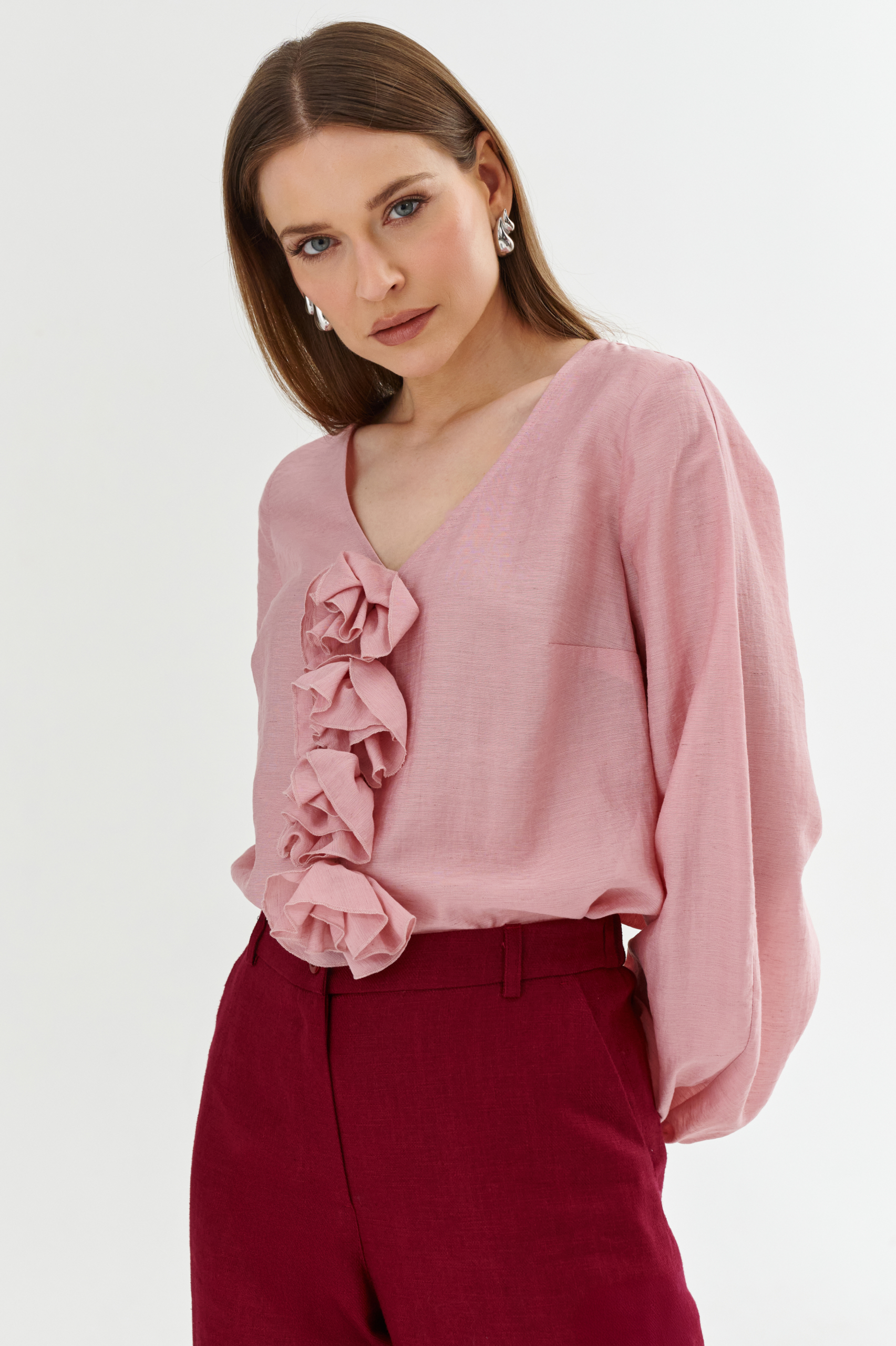 Różowa bluzka z dekoltem V z odpinanymi różami przypiętymi na agrafkę, długi rękaw, stylizacja z bordowymi spodniami z wysokim stanem.