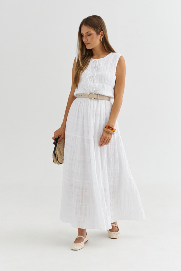 Modelka w białej sukience maxi z wiązaniami z przodu, fakturowana tkanina, stylizacja z beżowym paskiem.