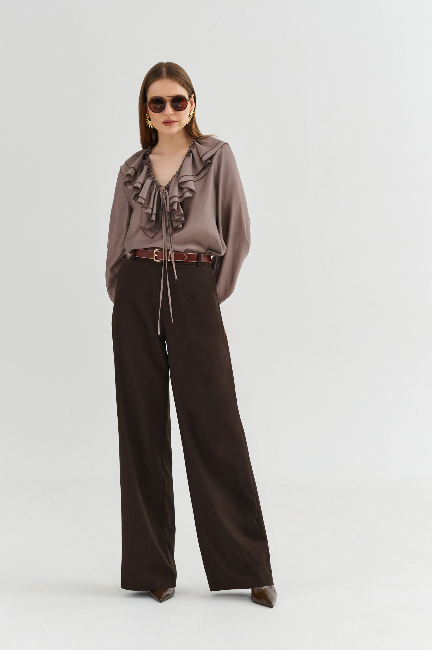 Modelka w bluzce z falbanami i wiązaniem przy dekolcie, z długim rękawem, zestawionej z brązowymi spodniami