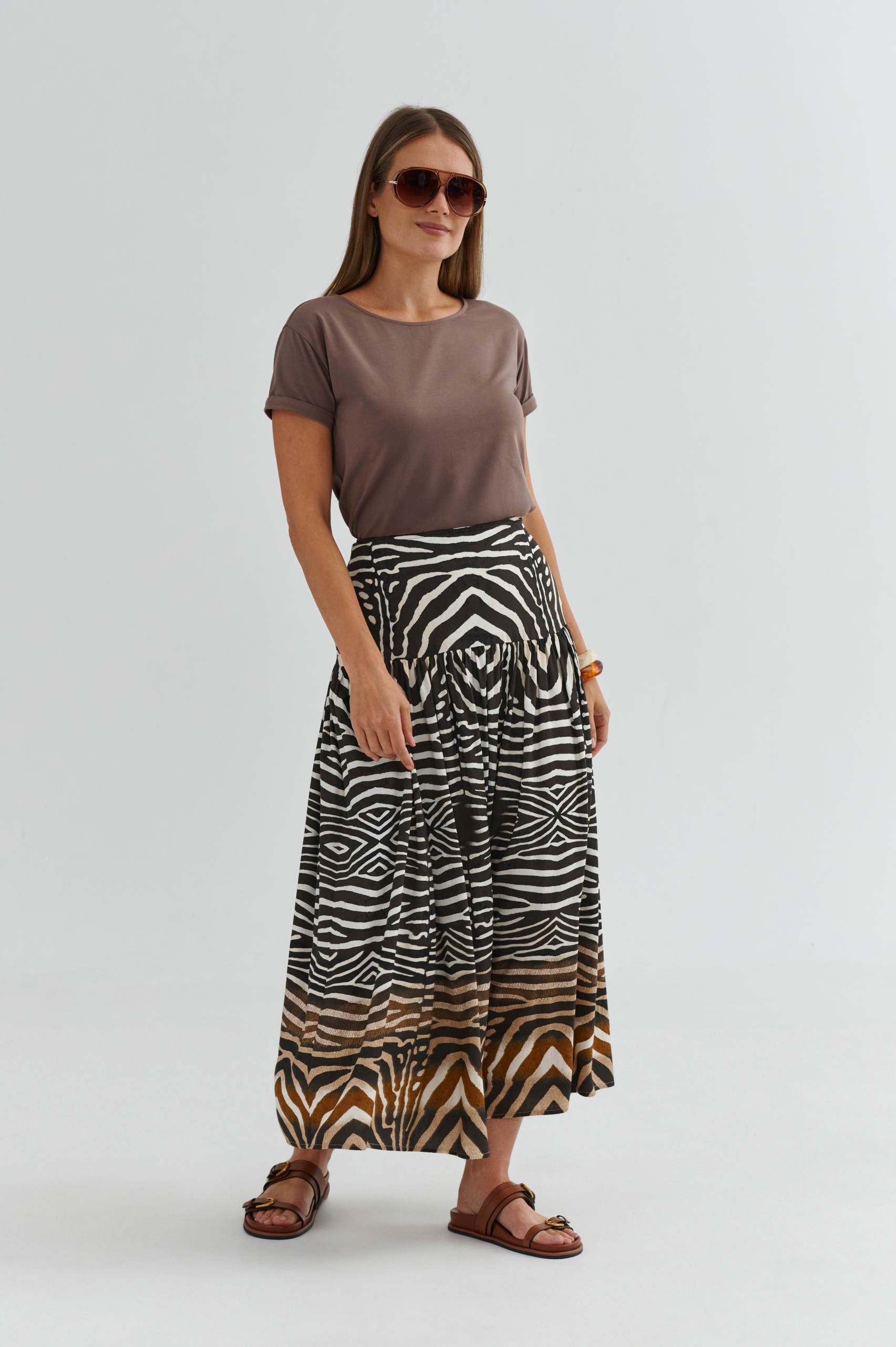 Modelka w t-shircie damskim w kolorze mokka z krótkim rękawem oraz spódnicy midi we wzór zebra, w brązowych sandałach i okularach.