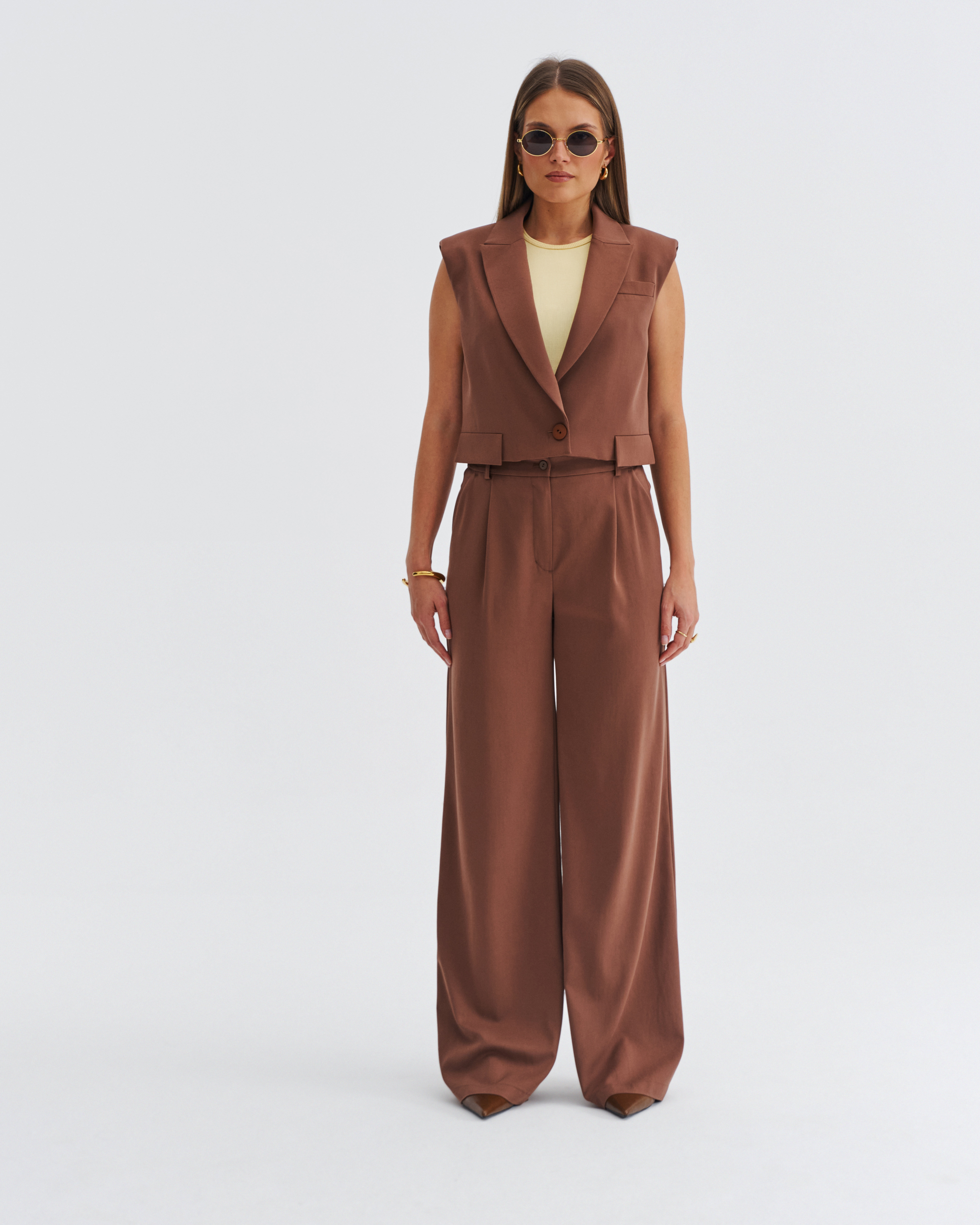 Brązowa damska kamizelka z szerokimi klapami i krótszym fasonem, noszona w komplecie z szerokimi spodniami.