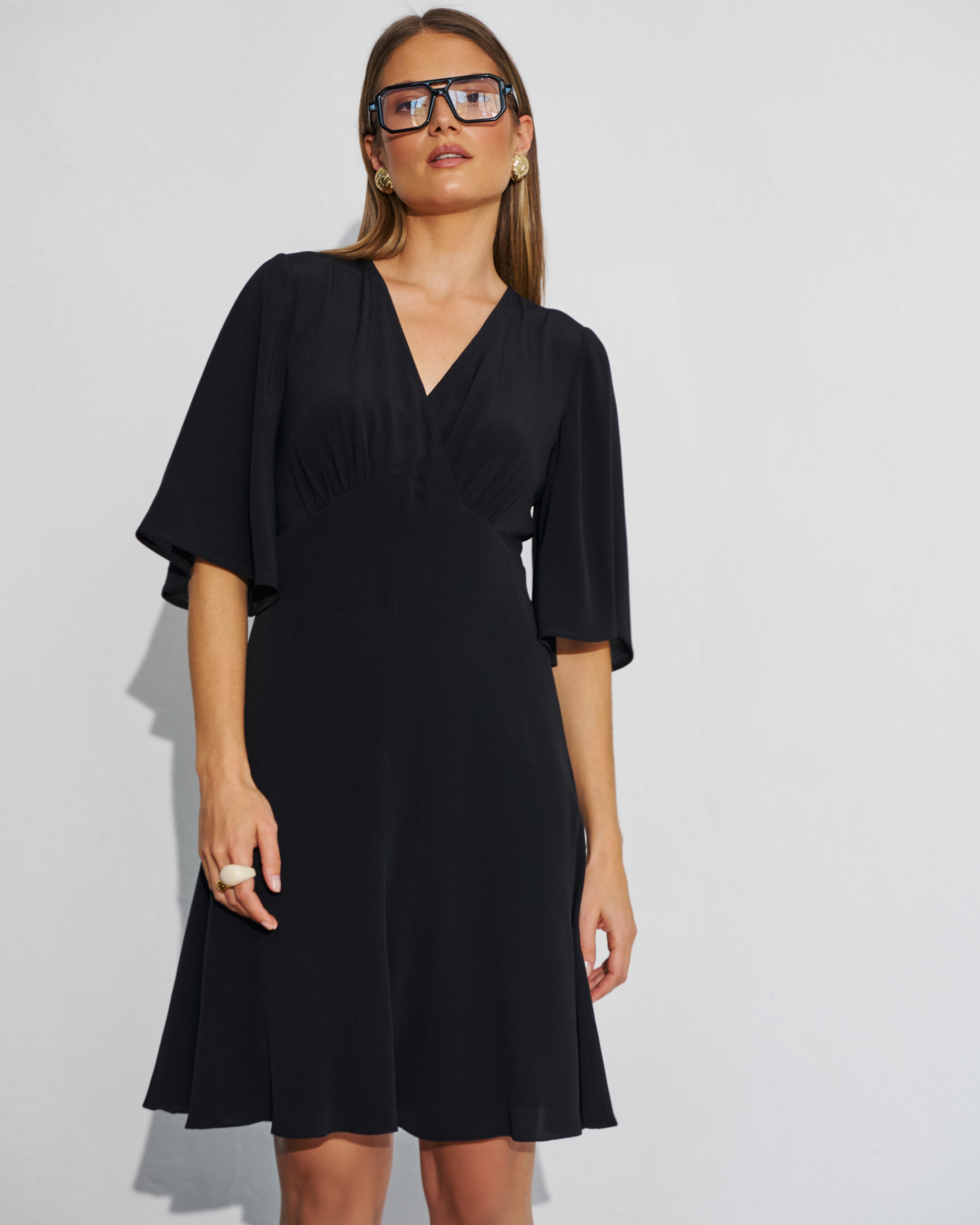 Modelka w czarnej krótkiej sukience rozkloszowanej z kopertowym dekoltem i szerokimi rękawami